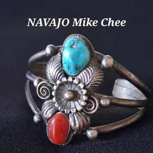 Puede incluir: Un brazalete de plata con piedras turquesa y coral. El brazalete está decorado con un diseño floral y lleva la firma de Navajo Mike Chee.