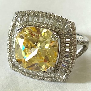 May include: A silver-toned ring featuring a large, cushion-cut yellow center stone. The stone is surrounded by a double halo of small, clear, baguette-cut and round-cut stones set in a square frame, with a split shank band design.
