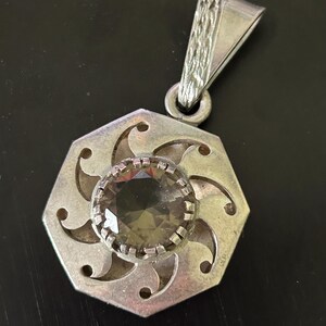 May include: A silver pendant with a large, round, smoky-colored gemstone at its center. The pendant has a geometric, floral-like design with cut-out details. The bail is triangular with a textured pattern.