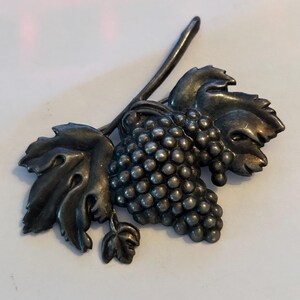 May include: A silver-toned brooch in the shape of a grape cluster with leaves and a stem. The brooch has a detailed design, with individual grapes and veined leaves. The metal has a slightly aged appearance.