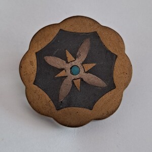May include: A gold-toned metal pin with a scalloped edge. The centre of the pin features a black background with a gold and turquoise starburst design.
