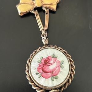 Peut inclure: Broche vintage dorée, avec un nœud en haut et un pendentif. Le pendentif présente un motif rond avec une rose rose et des feuilles vertes sur fond blanc. La broche a une bordure torsadée.
