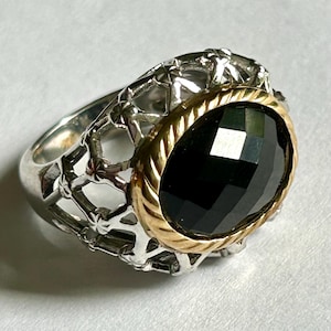 Pode incluir: Um anel de prata com uma faixa dourada e uma grande pedra preciosa preta facetada. O anel tem um design vazado na faixa, adicionando um elemento decorativo. A pedra preciosa é oval e capta a luz, criando um efeito brilhante.