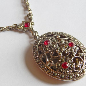 May include: A silver necklace with a large oval pendant. The pendant is made of silver and has a filigree design with red gemstones. The necklace is on a white background.