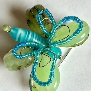 Könnte beinhalten: Eine grüne und blaue Schmetterlingsbrosche aus Glasperlen und Draht. Der Schmetterling hat einen hellblauen Körper und grüne Flügel mit blauen Perlenakzenten.