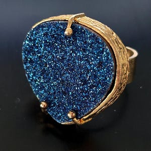 May include: A gold-toned ring featuring a large, heart-shaped, sparkling blue stone. The stone is set in a textured gold frame with a detailed band. The ring has a bold, eye-catching design.