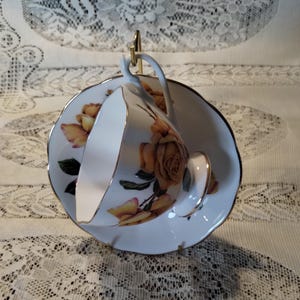 May include: White teacup and saucer set with a gold rim and a floral rose design in shades of yellow and orange. The teacup is suspended on a gold-colored metal stand, showcasing the delicate details of the porcelain.