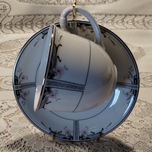 May include: A white teacup and saucer with a black and gray floral design. The cup and saucer have a gold trim.