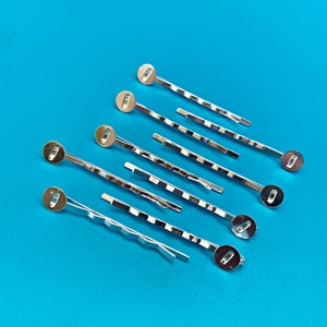 May include: A set of ten silver hair clips with round metal ends. The clips are arranged in a fan shape on a blue background.