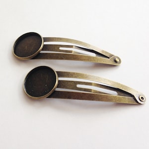 May include: Two antique bronze-colored hair barrettes. Each barrette has a flat, round base and a hinged, elongated clip. The clips are approximately 3 inches long, with a simple, elegant design.
