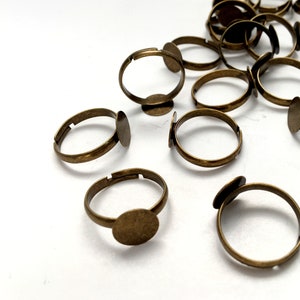 May include: Adjustable antique bronze-colored ring blanks with a flat, round pad for attaching embellishments. The rings are arranged on a white surface, showcasing their simple design and adjustable bands. Ideal for jewelry making and crafting.