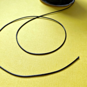 Pode incluir: Um cord&atilde;o el&aacute;stico preto enrolado em uma superf&iacute;cie amarela. O cord&atilde;o est&aacute; pronto para ser usado em projetos de artesanato.