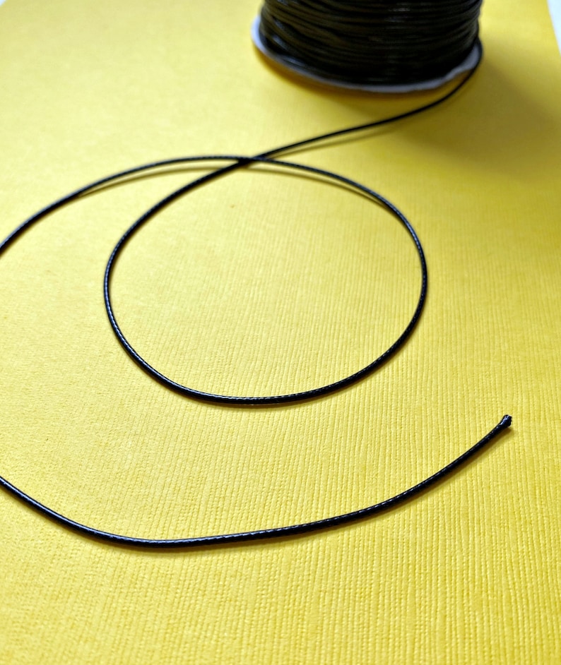 Pode incluir: Um cord&atilde;o el&aacute;stico preto enrolado em uma superf&iacute;cie amarela. O cord&atilde;o est&aacute; pronto para ser usado em projetos de artesanato.