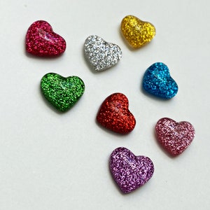 Op de afbeelding: Acht hartvormige glitterversieringen in verschillende kleuren, waaronder rood, groen, zilver, goud, blauw, roze en twee tinten rood.