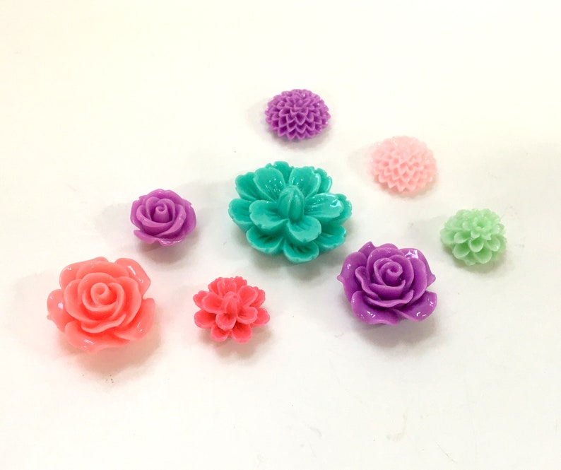 K&ouml;nnte beinhalten: Sortiment aus bunten Plastikblumenverzierungen in verschiedenen Formen und Gr&ouml;&szlig;en, darunter Rosen, G&auml;nsebl&uuml;mchen und Nelken. Die Blumen sind in den Farben Rosa, Lila, Gr&uuml;n und T&uuml;rkis.