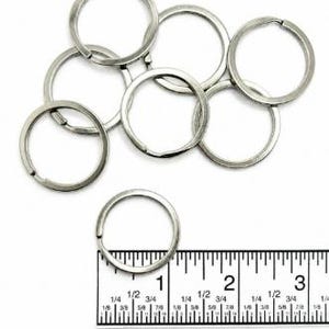 Può includere: Un insieme di sette anelli divisi in metallo color argento. Un anello è posizionato direttamente sopra un righello, mostrando un diametro di circa 2,54 cm. Gli anelli sono realizzati in filo metallico avvolto per contenere chiavi.