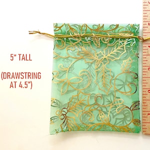 K&ouml;nnte beinhalten: Eine gr&uuml;ne Organza-Kordeltasche mit goldenem Blumenmuster. Die Tasche ist 12,7 cm hoch und hat einen Kordelzug bei 11,4 cm.