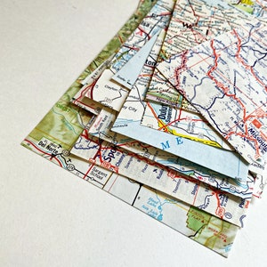 May include: A stack of vintage paper maps with various locations and roads marked in blue, red, and black. The maps are folded and layered, creating a textured and colorful composition.