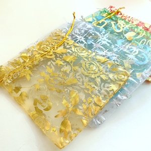 K&ouml;nnte beinhalten: Drei durchsichtige Organza-Geschenkbeutel mit goldenen Blumenmustern. Die Beutel liegen &uuml;bereinander, der goldene Blumenbeutel unten, ein hellblauer Beutel in der Mitte und ein silberner Beutel oben.