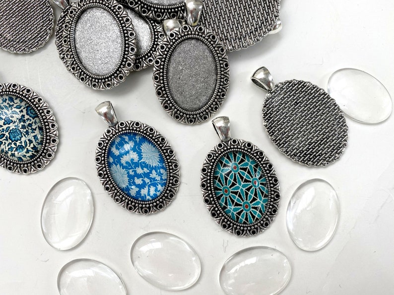 Puede incluir: Surtido de bandejas ovaladas de plata para colgantes con dise&ntilde;os ornamentados. Algunas bandejas est&aacute;n vac&iacute;as, mientras que otras presentan insertos de vidrio con patrones coloridos. Tambi&eacute;n se incluyen varios cabujones de vidrio transparente.