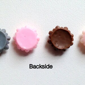 K&ouml;nnte beinhalten: Vier runde, dekorative Kn&ouml;pfe mit einem gewellten Rand. Die Kn&ouml;pfe sind grau, rosa, braun und rosa. Die Kn&ouml;pfe sind von links nach rechts in einer Reihe angeordnet. Der Text "Backside" ist unter den Kn&ouml;pfen gedruckt.
