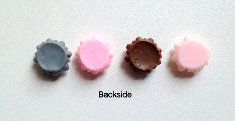 K&ouml;nnte beinhalten: Vier runde, dekorative Kn&ouml;pfe mit einem gewellten Rand. Die Kn&ouml;pfe sind grau, rosa, braun und rosa. Die Kn&ouml;pfe sind von links nach rechts in einer Reihe angeordnet. Der Text "Backside" ist unter den Kn&ouml;pfen gedruckt.