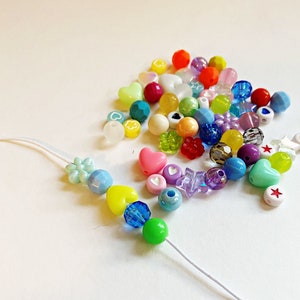 May include: A colorful assortment of plastic beads in various shapes, including hearts, stars, and flowers. The beads are strung on a white cord, creating a necklace or bracelet.