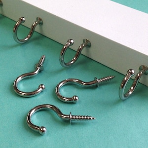 May include: Six silver metal hooks with screws. The hooks are in a u-shape and have a rounded end. The hooks are on a white surface with a turquoise background.