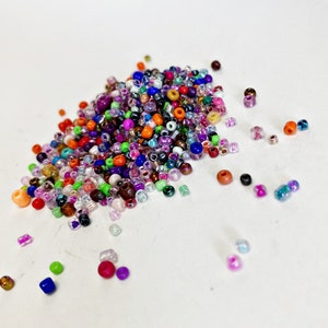 May include: A large pile of colorful seed beads in various shades of purple, blue, green, red, orange, brown, and white. The beads are scattered on a white surface.