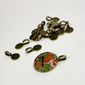 May include: A pile of 18 antique gold-toned metal pendant bails, and a single oval-shaped pendant with a colourful floral design.