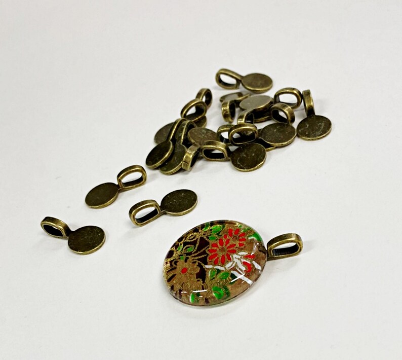 May include: A pile of 18 antique gold-toned metal pendant bails, and a single oval-shaped pendant with a colourful floral design.