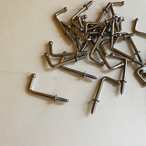 May include: A pile of silver metal hooks with screws. The hooks are all the same size and shape, and they are arranged in a random pattern.