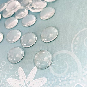 K&ouml;nnte beinhalten: Eine Sammlung von transparenten Glascabochons, auch bekannt als Glasperlen, auf einem hellblauen Hintergrund mit einem wei&szlig;en Blumenmuster.