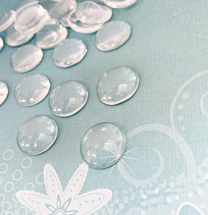 K&ouml;nnte beinhalten: Eine Sammlung von transparenten Glascabochons, auch bekannt als Glasperlen, auf einem hellblauen Hintergrund mit einem wei&szlig;en Blumenmuster.