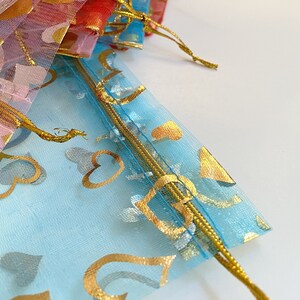 K&ouml;nnte beinhalten: Eine hellblaue, durchsichtige Tasche mit goldenen Herzmustern und einem goldenen Kordelzug. Die Tasche ist aus einem zarten, durchsichtigen Material gefertigt.