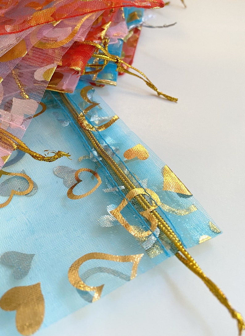 K&ouml;nnte beinhalten: Eine hellblaue, durchsichtige Tasche mit goldenen Herzmustern und einem goldenen Kordelzug. Die Tasche ist aus einem zarten, durchsichtigen Material gefertigt.