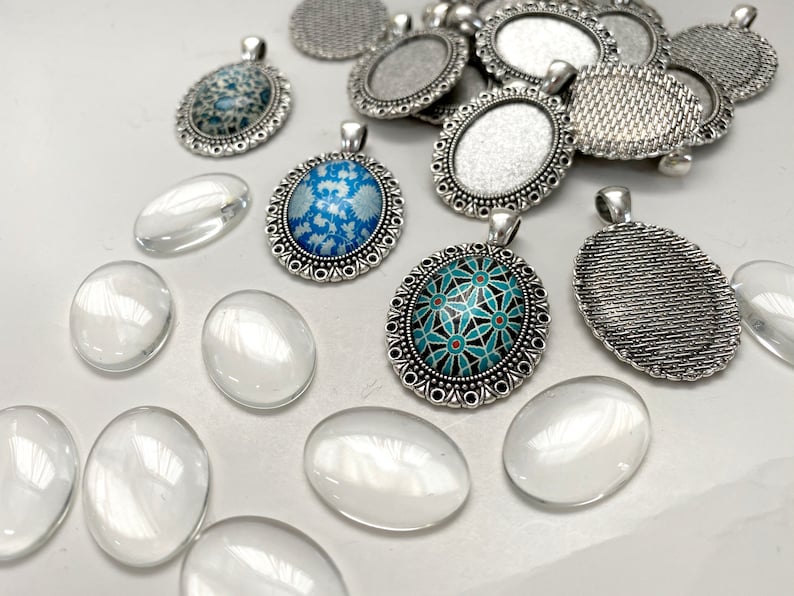 Puede incluir: Surtido de bandejas ovaladas de plata para colgantes con bordes decorativos, algunas con inserciones de vidrio con patrones azules y turquesa, y cabujones de vidrio transparentes.