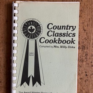 May include: A spiral-bound cookbook titled "Country Classics Cookbook" with a light green cover. The cover features a black and white ribbon design with a maple leaf emblem and the text "Judge's Special". The text "The Award Winning Recipes From Local Fall Fairs" is also present.