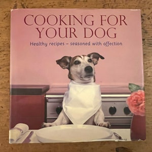 May include: A book cover with a white background and the title "Cooking for Your Dog" in red text. The subtitle is "Healthy recipes - seasoned with affection". A dog wearing a white bib is sitting at a table with a plate in front of it.