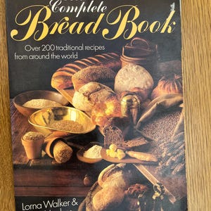 May include: The cover of "The Complete Bread Book" features a collection of various breads, grains, and ingredients. The title is in elegant script, with the subtitle "Over 200 traditional recipes from around the world." Authors Lorna Walker & Joyce Hughes are listed.