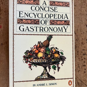 May include: A white book with a black and brown illustration of a bowl of fruit on the cover. The title is "A Concise Encyclopedia of Gastronomy" by Andre L. Simon.