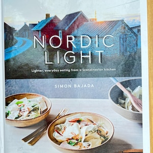 May include: The cookbook "Nordic Light" by Simon Bajada, with the subtitle "Lighter, everyday eating from a Scandinavian kitchen". The cover shows a landscape with red buildings and a photo of food in bowls.