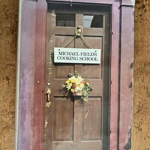 Puede incluir: Una portada de libro vintage con una foto de una puerta de madera con un letrero que dice "Michael Field's Cooking School". Un arreglo floral está colgado en la puerta.