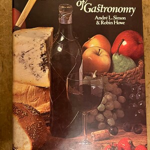 May include: A book titled "Dictionary of Gastronomy" by Andre L. Simon & Robin Howe. The cover features a still life of a glass of red wine, a bottle of red wine, a loaf of bread, a wedge of blue cheese, grapes, apples, a pear, and strawberries.
