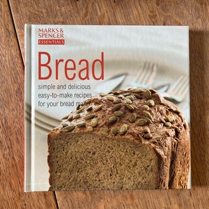 May include: A cookbook titled "Bread: simple and delicious easy-to-make recipes for your bread maker" by Marks & Spencer Essentials, featuring a close-up image of a loaf of bread with pumpkin seeds.