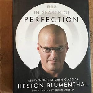 May include: A cookbook titled "In Search of Perfection" by Heston Blumenthal. The cover shows a portrait of the author in a white circle against a black background. The book is about reinventing kitchen classics.