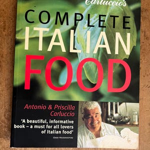 May include: A book titled "Carluccio's Complete Italian Food" by Antonio & Priscilla Carluccio. The cover features a green leafy background with a red title and a photo of a man smiling. The quote "A beautiful, informative book - a must for all lovers of Italian food" is printed below the photo, attributed to Good Housekeeping.