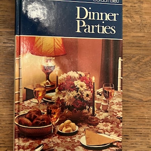 May include: A cookbook titled "Dinner Parties" by Cordon Bleu, featuring a photo of a beautifully set table with a floral centrepiece, candles, wine glasses, and bread rolls.