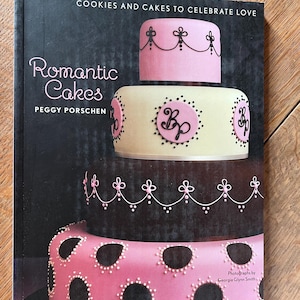 May include: A book titled "Romantic Cakes: Cookies and Cakes to Celebrate Love" by Peggy Porschen. The cover features a multi-tiered wedding cake decorated with pink and black fondant, intricate piping, and the initials "B.P."