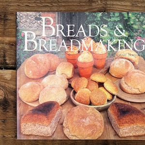 May include: A cookbook titled "Breads & Breadmaking" with various baked goods. The image features an assortment of breads, rolls, and baked goods on a wooden surface. The book cover displays the title in large, elegant lettering.